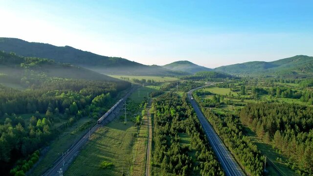 The movement of trains and trucks parallel to each other in a picturesque place. Railway tracks and a highway in the middle of the forest and mountains.
