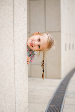 A blonde girl with a pigtail sticks her head out from behind a pillar