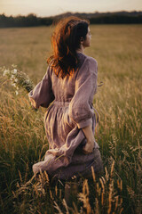 Beautiful woman in linen dress running with wildflowers in hand in summer meadow in sunset. Stylish...