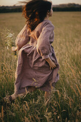 Beautiful woman in linen dress running with wildflowers in hand in summer meadow in sunset....