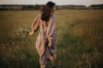Beautiful woman in linen dress running with wildflowers in hand in summer meadow in sunset....