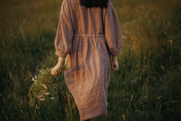 Beautiful woman in linen dress walking with wildflowers in hand in summer meadow in sunset, back...