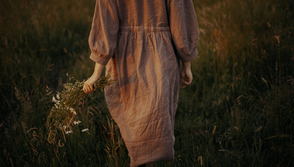 Beautiful woman in linen dress walking with wildflowers in hand in summer meadow in sunset....