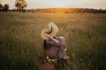 Obraz premium Beautiful woman in linen dress and straw hat sitting on rustic chair in summer meadow in sunset. Young female with basket of flowers relaxing in countryside. Atmospheric calm tranquil moment