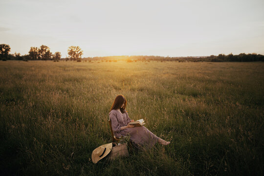 Beautiful Woman In Linen Dress Reading Book In Summer Meadow In Sunset. Young Female With Book And Basket Of Flowers Sitting On Rustic Chair And Relaxing In Countryside. Atmospheric Calm Moment