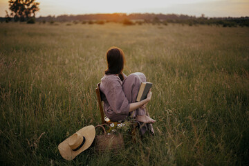 Beautiful woman in linen dress sitting on rustic chair and enjoying sunset in summer meadow. Young...