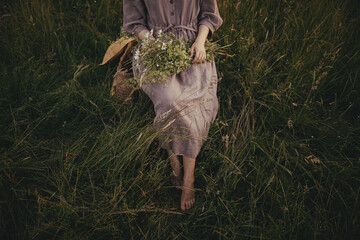 Beautiful woman in linen dress sitting barefoot on rustic chair among grass with bouquet of...