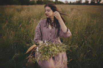 Beautiful woman with windy hair and in linen dress sitting on wooden chair with bouquet of flowers...