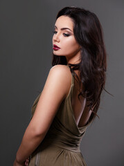 Naklejka premium Beautiful bright makeup woman with long black curly volume black hair style in green elegant dress with nude back, burgundy lipstick looking on grey background.. Closeup