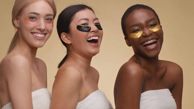 Close up portrait of three young diverse ladies with collagen eye patches laughing to camera, posing wrapped in towels
