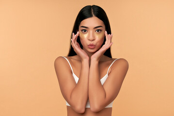 Fototapeta premium Young Asian woman in white lingerie and clean radiant skin with moisturizing patches under the eyes on a beige background. Spa care, cosmetology.