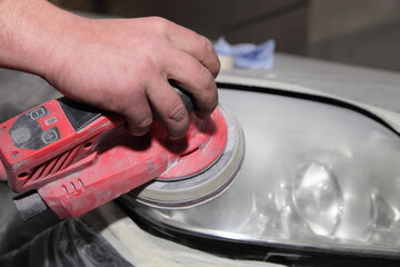 Car headlamp abrasive grind with rotary grinding machine before buffing in serviceman hand closeup, vehicle body renovation technology