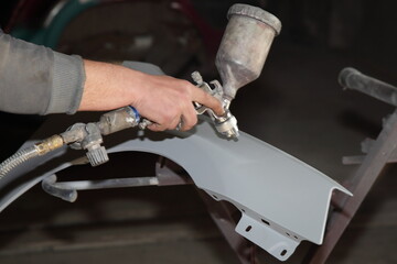 Service man hand paints a car body part new fender with ground painting gun on holding frame device, vehicle body repair service