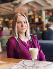 A blonde girl in a lilac sweater sits in a cozy restaurant on a sofa and she is relaxed and smiling and have a coffee drink on her table.