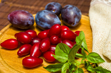 Still life - fresh large dogwood berries, purple plums and a sprig of basil on a wooden board