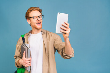 Portrait of happy smiling young student standing with backpack and folders isolated over blue background. Using tablet.