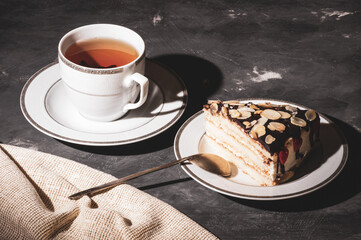 Still life - layered homemade sponge cake and a cup of black tea in a white dish, and a linen napkin, black background, hard light, photo in a low key.