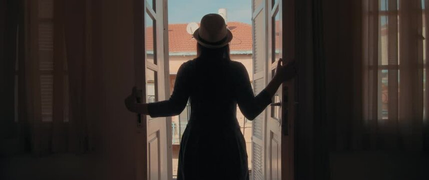 A woman in a hat opening old glass doors of the apartment to go out to balcony and enjoy the view of old town. ravel, european summer vacation. Slow motion, handheld. 
