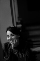 A happy young girl in a black leather jacket and short hair is sitting at a table. Black and white portrait of a smiling woman