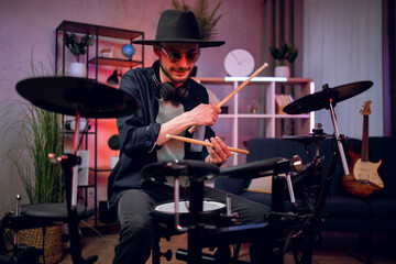 Fototapeta premium Happy stylish man sitting at dark music studio and playing electronic drums with sticks. Professional musician wearing trendy black hat and sunglasses.