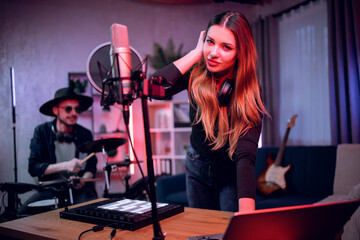 Charming woman in headphones using laptop, microphone and dj controller for creating music in studio. Handsome man in hat and glasses playing electronic drums on background.