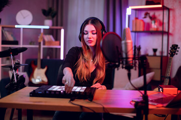 Beautiful female dj in headphones mixing sounds on laptop and digital controller. Young woman sitting at dark atmosphere and creating modern music.