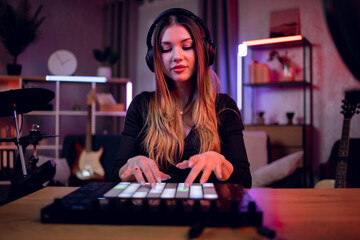 Inspired woman in headphones sitting in dark atmosphere and making music with help of modern dj controller. Creative process at recording studio.