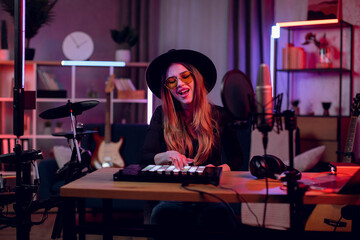 Female musician in trendy hat and glasses singing in microphone and using mixer console while...