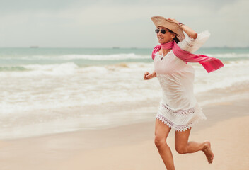 middle-aged woman in white dress and pamela runs along the shore of the beach happy. enjoying the beach. vacation and maturity concept. healthy middle age.