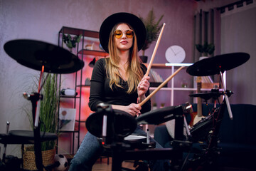 Beautiful young woman in black hat and eyeglasses playing electronic drums indoors. Female musician...