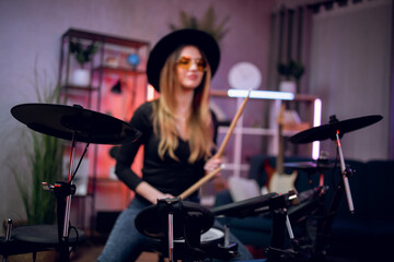 Fototapeta na wymiar Inspired young lady in stylish outfit using electronic drums while recording sound at studio. Charming woman playing on music instrument at dark atmosphere,
