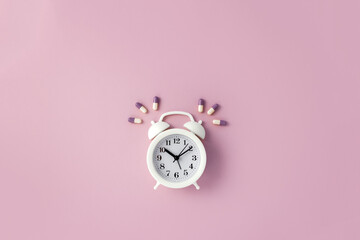 Alarm clock surrounded by colored pills on pastel pink background. Time to take vitamins concept....