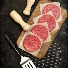 Ground Beef Patties for Grilling and Roasting. Raw Minced Steak Burgers from Beef Meat on Black Background. BBQ Grill Tools with Raw Beef Hamburger Patties. Burger Cutlets On Paper And Grill Grate.