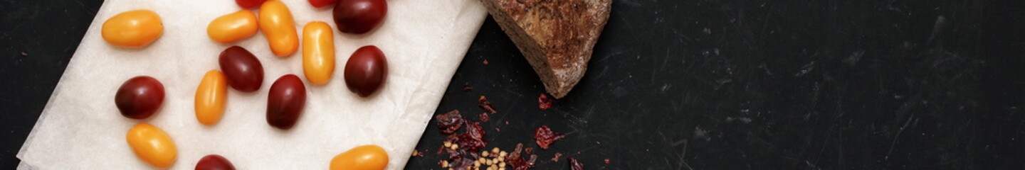 Plum Cherry Tomatoes On Culinary Paper, Bread And Dried Chili Peppers. Scattered Red, Yellow And Black Mini Cherry Tomato On Black Grunge Background.