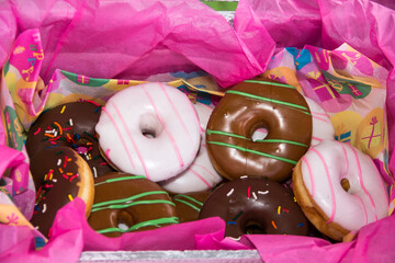 Box with a bulk of different type of colorful tasty gluten free doughnut surround by pink wrapping paper
