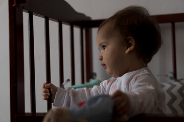 Cute cheerful little chubby baby girl sitting in grey baby crib after waking up from sleep in white and pink pajamas with teddy bear and smiling. Childhood, leisure, comfort, medicine, health concept