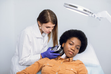 Young African American woman client lying on the couch in modern dermatology clinic, having face...