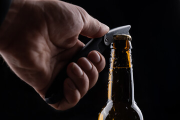 A man opens a bottle of beer from a dark glass with drops of condensation on a black background.