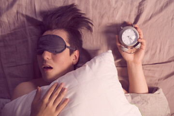 A young funny man sleeps soundly early in the morning, hugs a pillow and holds a vintage alarm...