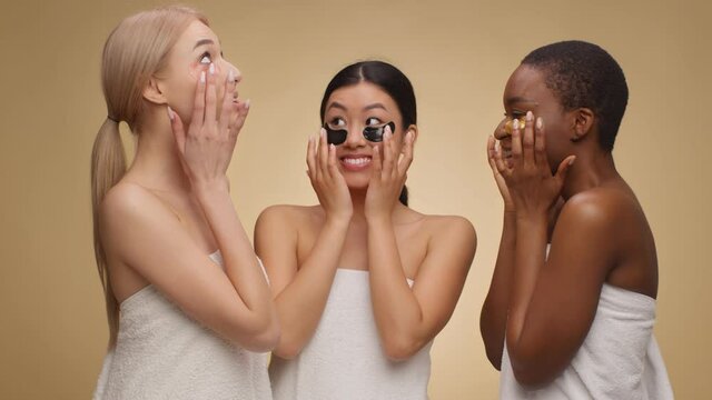 Positive diverse women wrapped in towels putting gel patches on under eyes area and laughing, beige studio background