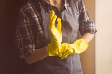 A young girl in a plaid shirt and apron puts yellow rubber gloves on her hands to start cleaning her house and create comfort. Housekeeper with gloves doing disinfection.