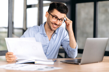 Happy successful confident young caucasian businessman, manager or freelancer wearing glasses sitting at table in office enjoying profit, holding business documents, looking at laptop screen, smiling