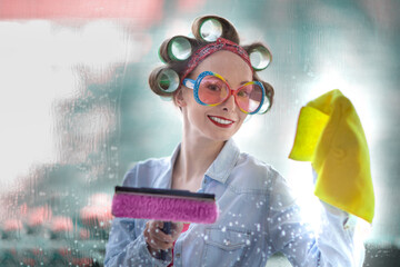 Young woman with curlers in her hair cleans a window with rag and wiper in hand