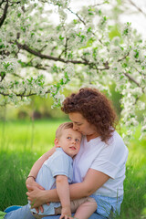 mother sit, holds young son in flowering spring garden. parental love and care