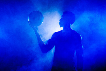 Basketball player holding a ball against blue fog background. African american man silhouette