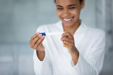 Excited young mixed race pregnant woman happy about positive test result, expecting baby, getting pregnancy confirmation, looking at tester in bathroom, smiling, laughing. Fertility concept. Close up