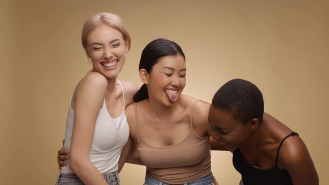 Three happy multiethnic friends laughing together, grimacing and sticking out tongues, beige background, slow motion