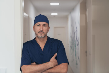 Portrait Of Mature Male Doctor In Busy Modern Hospital Corridor. 