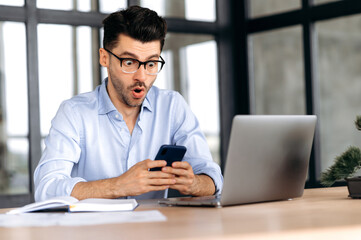Fototapeta Excited surprised caucasian young broker, manager or freelancer wearing glasses, sitting at his desk in the office, using smartphone, shocked by news or message, looking at screen in amazement obraz