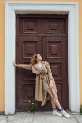 Young smiling millennial woman with wild hair dressed in an autumn coat posing near the door of an old building.